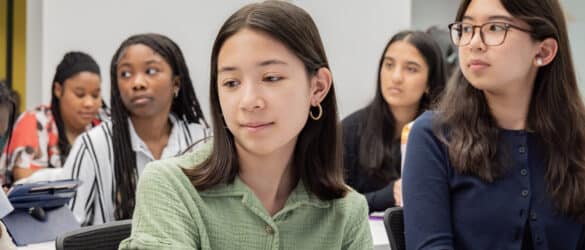 A group of students sit in a classroom, listening attentively. The focus is on a girl in a green shirt in the foreground, with several other students visible behind her.