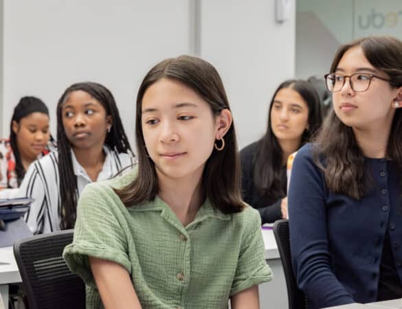 A group of students sit in a classroom, listening attentively. The focus is on a girl in a green shirt in the foreground, with several other students visible behind her.