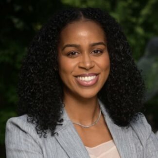 A woman with curly black hair, wearing a light gray blazer and necklace, smiles at the camera with greenery in the blurred background.