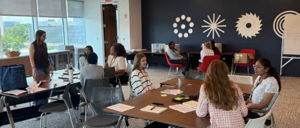 Several people sit and talk around tables in a modern meeting room with large windows, abstract wall art, and scattered papers. One person stands and addresses the group. The atmosphere is casual and collaborative.