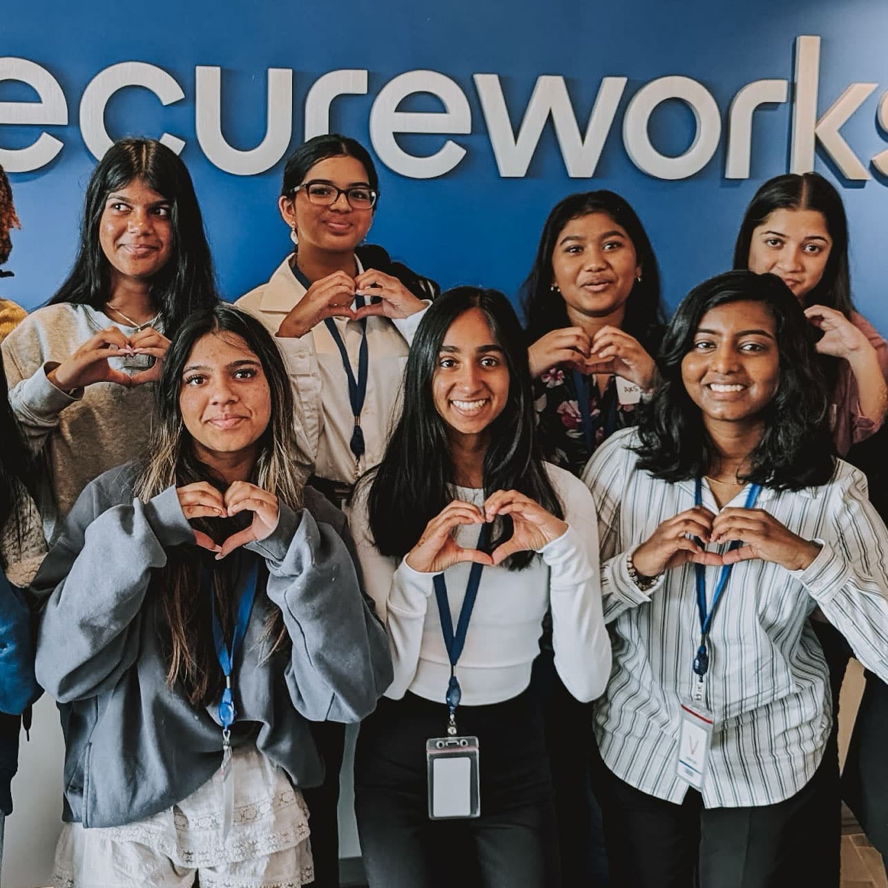 A group of smiling women, wearing lanyards, make heart shapes with their hands in front of a blue wall with the word Secureworks partially visible in the background.