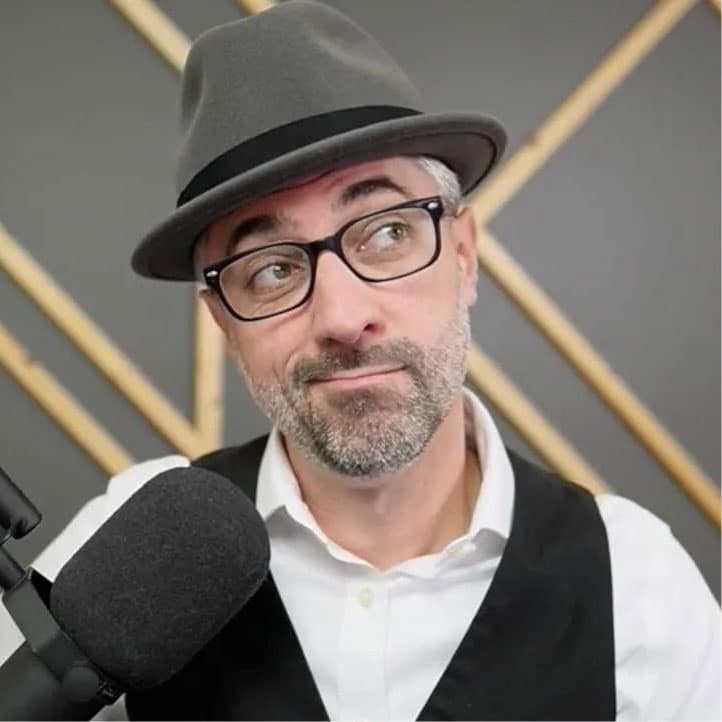 A man with glasses, a gray hat, and a beard looks to the side while sitting in front of a microphone. He wears a white shirt and black vest, with a geometric background behind him.