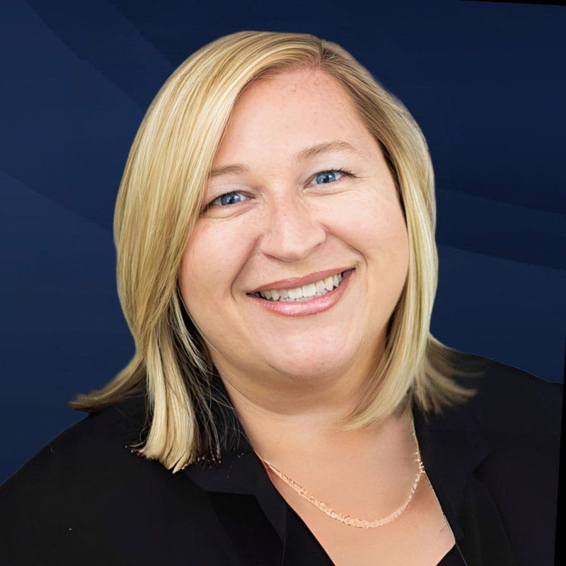 Smiling woman with straight blonde hair, wearing a black blazer and a thin gold necklace, poses in front of a dark blue background.