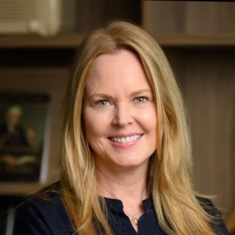 Woman with long blonde hair smiling, wearing a dark top and a necklace, standing indoors in front of wooden bookshelves with blurred objects and a framed picture in the background.