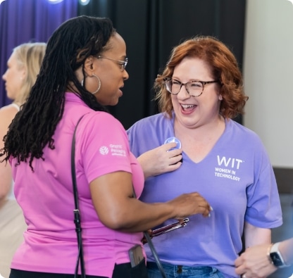 Two women smiling and talking at an event, one wearing a pink shirt and the other in a purple Women in Technology t-shirt with a name tag. Other people are blurred in the background.