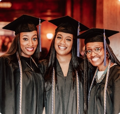Three women wearing graduation caps and gowns stand close together, smiling at the camera in a warmly lit indoor setting.
