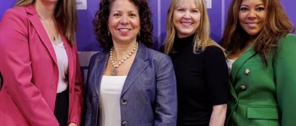 Four women stand together, smiling, in front of a purple Women in Technology backdrop. They are dressed in business attire: pink blazer, gray jacket, black top, and green suit.