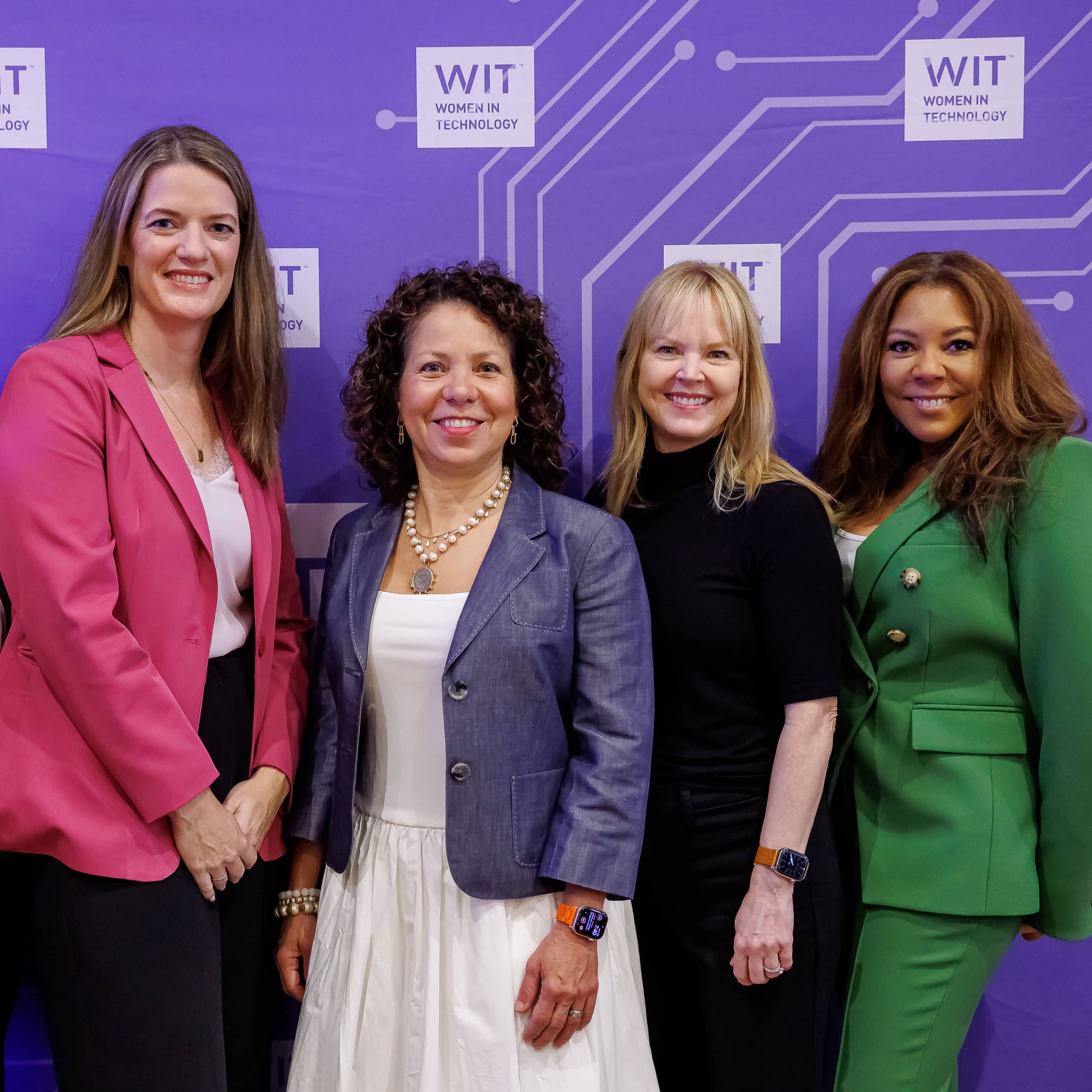 Four women stand together, smiling, in front of a purple Women in Technology backdrop. They are dressed in business attire: pink blazer, gray jacket, black top, and green suit.