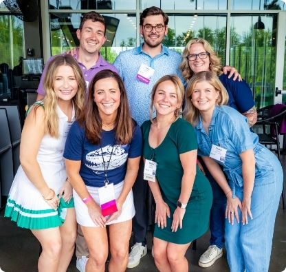 Seven adults stand together indoors, smiling at the camera. Three are kneeling in front, and four are standing behind. They wear casual and business casual outfits, and nametags. Large windows and greenery are visible in the background.