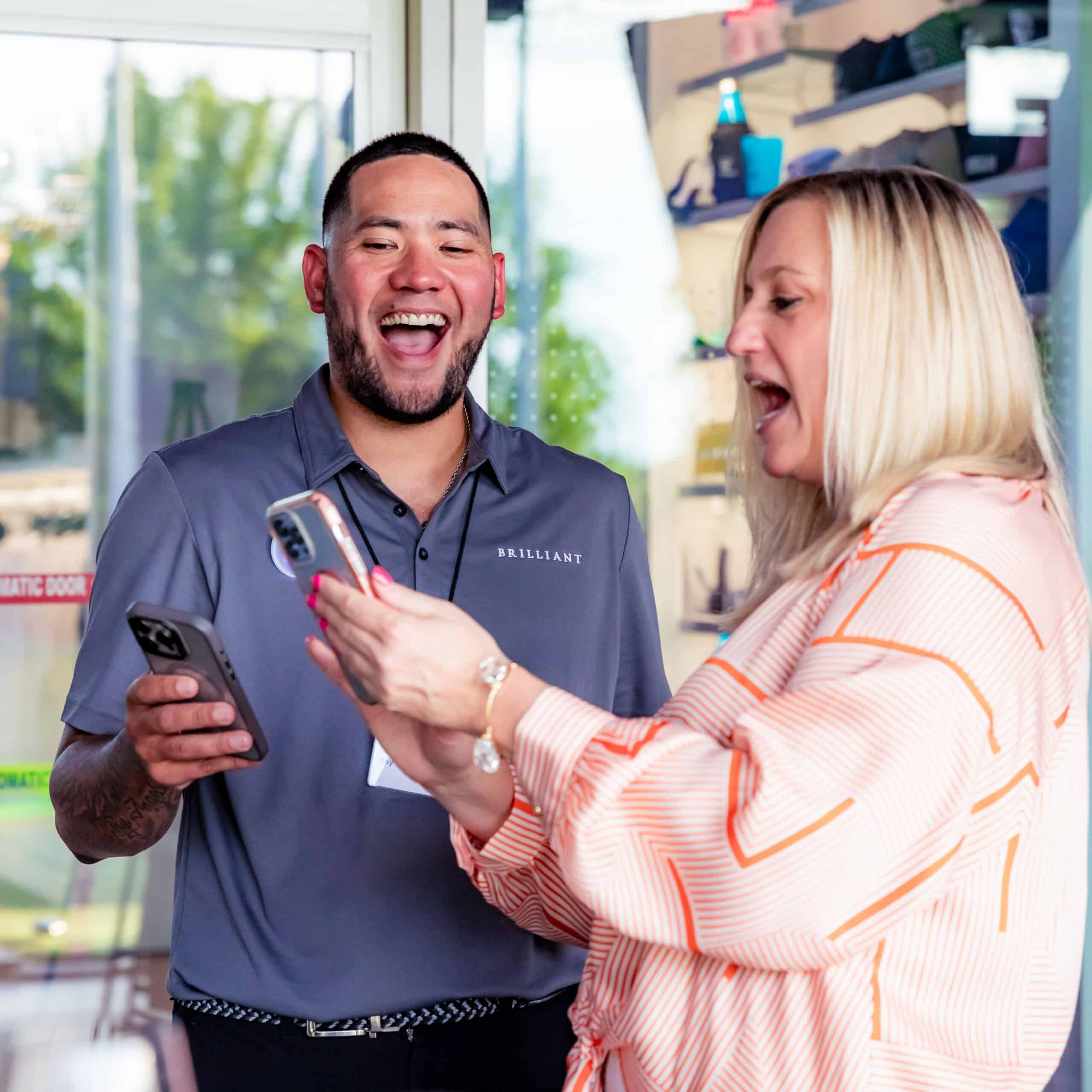 Two people smiling and laughing as they look at their phones together indoors. The man wears a gray polo shirt, and the woman wears an orange-striped blouse. Bright light comes through the windows behind them.