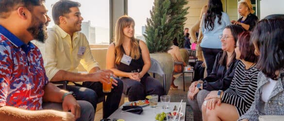 A group of people sit and smile together outdoors at a rooftop gathering, with food and drinks on the table in front of them. Others socialize in the background near a railing.