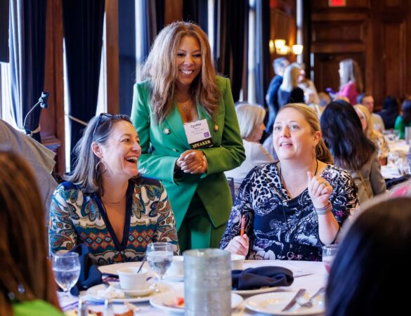 A woman in a green suit, wearing a speaker badge, smiles while standing among seated women who are talking and laughing at a round dining table in a conference or banquet setting.