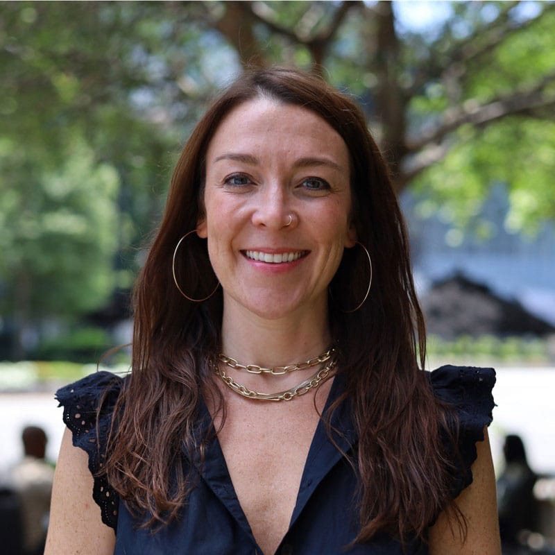 A woman with long brown hair, hoop earrings, and layered necklaces smiles outdoors. She wears a sleeveless black top, with trees and blurred greenery in the background.