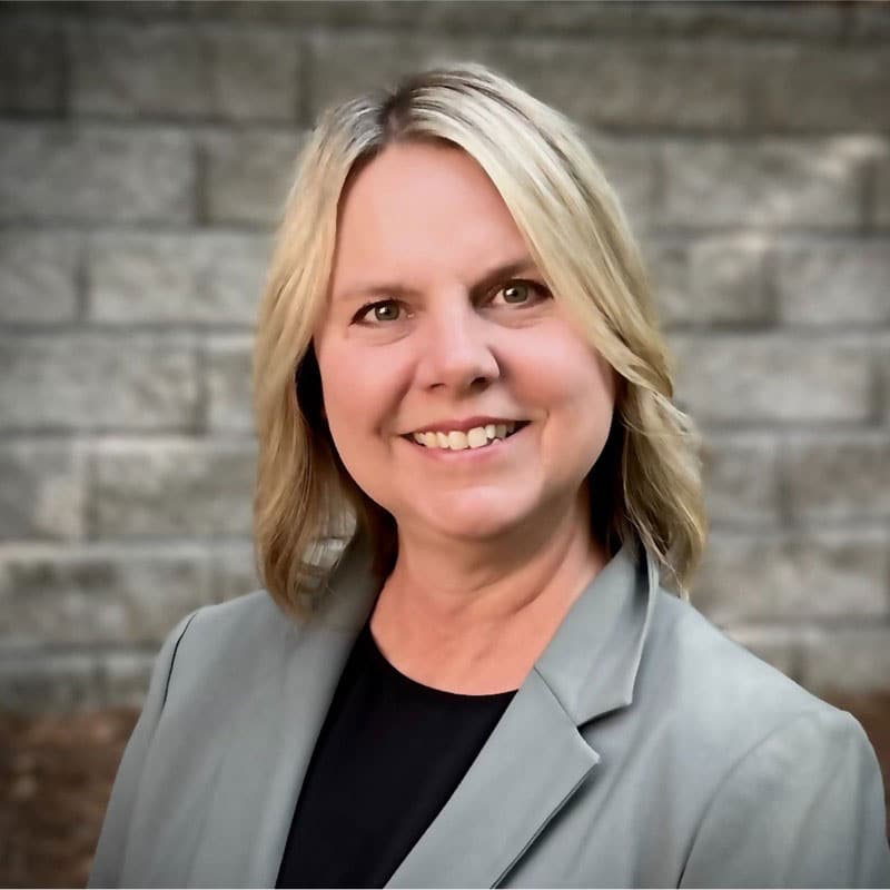 Smiling woman with shoulder-length blonde hair wearing a light gray blazer and black top, standing outside in front of a gray brick wall.