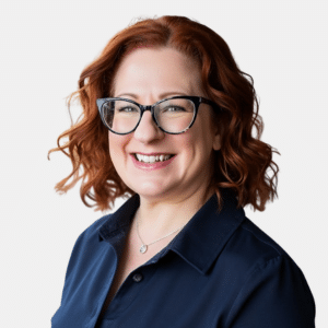 Smiling woman with curly red hair and glasses, wearing a navy collared shirt and a silver necklace, against a plain white background.