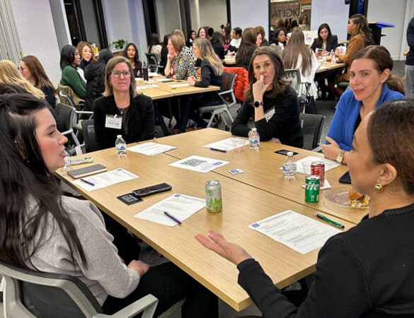 Five women sit around a table in discussion at a professional event, with papers, drinks, and name tags visible. Other groups are talking at nearby tables in a modern, well-lit room.