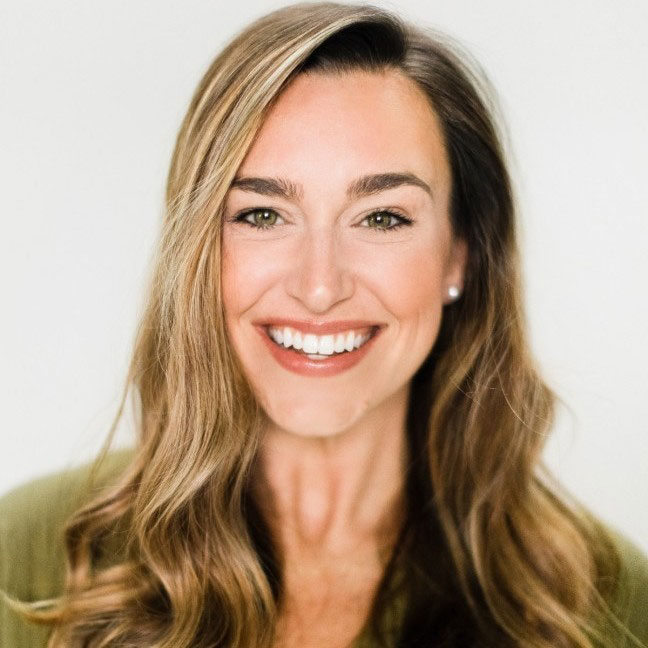 Smiling woman with long, wavy light brown hair and fair skin, wearing a green top, against a plain light background.