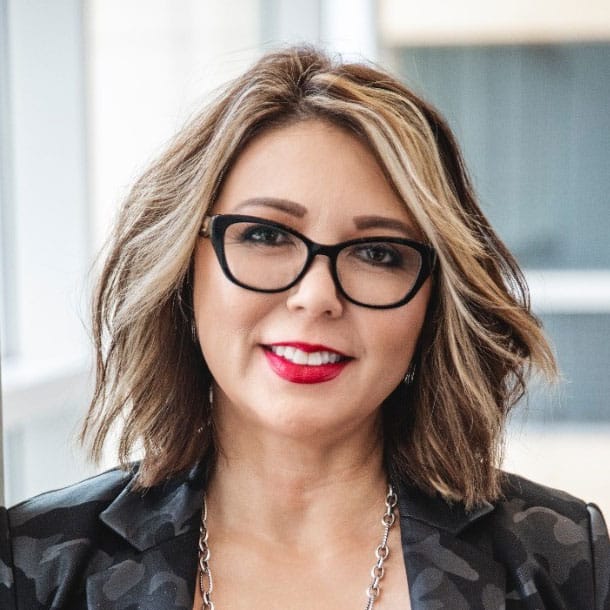 A woman with shoulder-length wavy hair, wearing glasses, a black patterned jacket, a necklace, and red lipstick, smiles at the camera in a bright indoor setting.