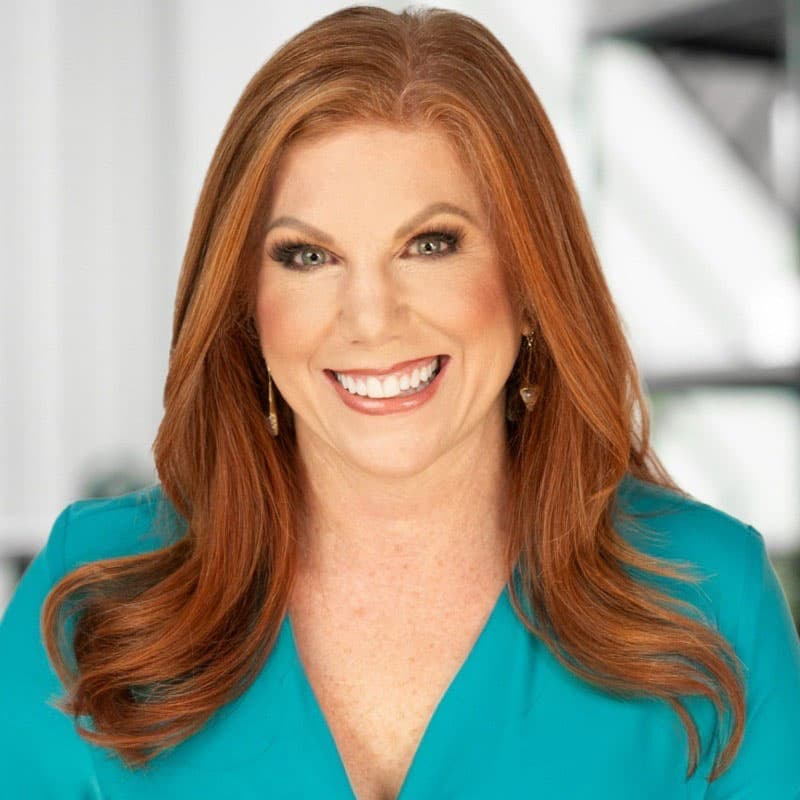 A woman with long red hair, wearing a turquoise top and gold earrings, smiles at the camera against a blurred indoor background.