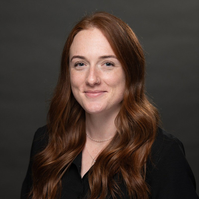 A woman with long wavy auburn hair wearing a black shirt and a delicate necklace smiles softly against a plain dark background.