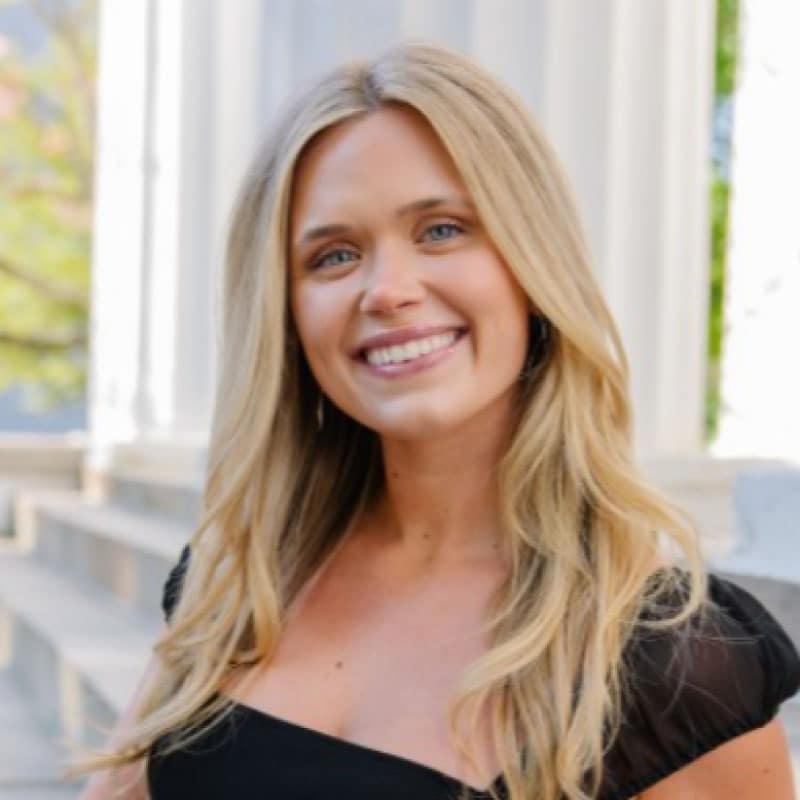 A young woman with long blonde hair and a black top smiles outdoors in front of white columns and stone steps.