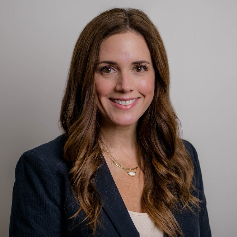 A woman with long brown hair, wearing a dark blazer and layered necklaces, smiles at the camera against a plain light background.
