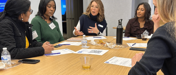 Five women sit around a wooden table, having a discussion. Papers, snacks, bottled water, and a black thermos are on the table in a modern, well-lit room.