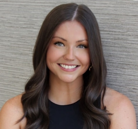 A woman with long brown hair and blue eyes smiles at the camera, wearing a sleeveless black top, seated in front of a textured gray background.