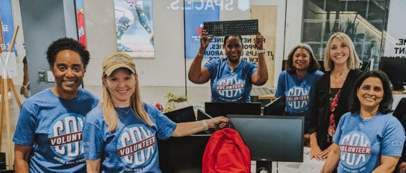 Six volunteers in matching blue shirts smile in a tech workspace, with one person holding a keyboard overhead and another holding a red backpack. Computer equipment and a glass wall are visible in the background.