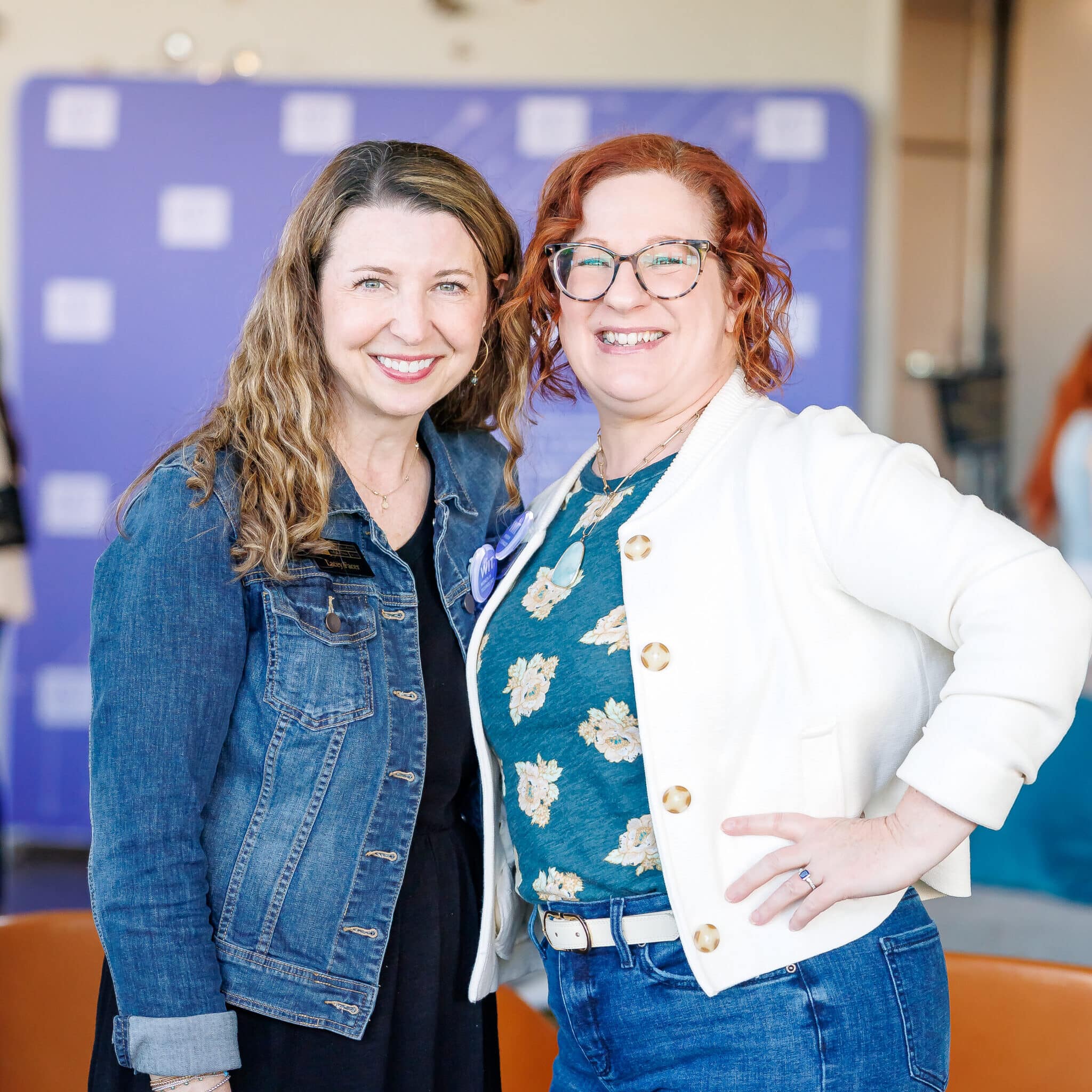 Two smiling women pose together indoors, one in a denim jacket and the other in a white jacket with gold buttons, standing in front of a purple backdrop.