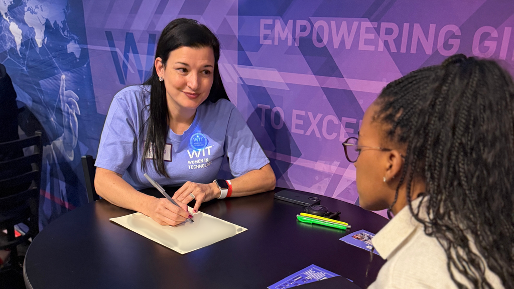 A woman in a WIT t-shirt talks with another woman at a table, writing on a paper. Highlighters, a phone, and pamphlets are on the table. The background reads EMPOWERING GIRLS TO EXCEL..