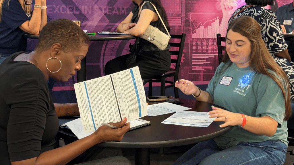 Two women sit at a round table reviewing documents together. One reads a paper intently while the other gestures, with more people and a colorful EXCEL IN STEAM banner in the background.