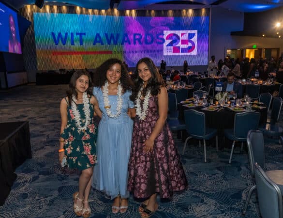 Three girls in floral dresses and leis smile together at an indoor event. A large screen in the background reads WIT AWARDS 25. Tables and guests are visible behind them in a dimly lit, elegant venue.