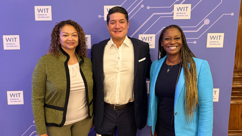 Three people stand smiling in front of a purple Women in Technology (WIT) backdrop, wearing business attire. Two women flank one man in the center.