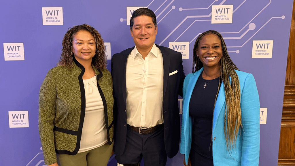Three people pose and smile in front of a purple backdrop with WIT Women in Technology logos. The group includes two women and one man dressed in business attire.