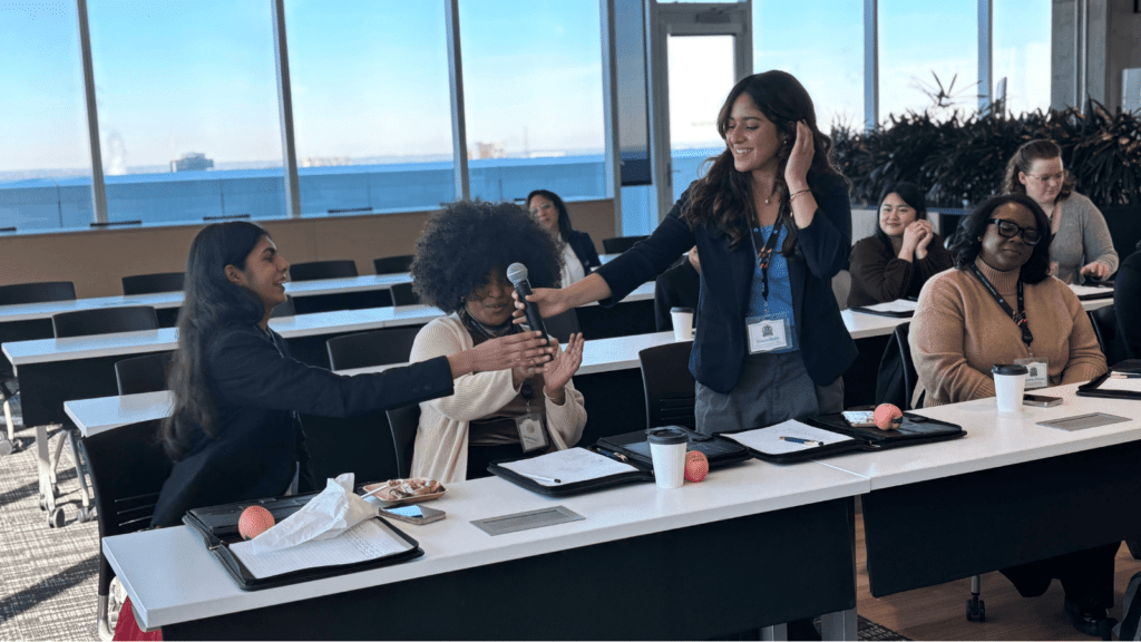 A woman hands a microphone to a smiling attendee in a conference room while others watch. The attendees have name badges, trays, and coffee cups on their desks.