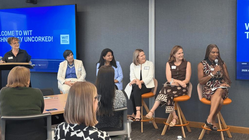 Five women sit on stools speaking at a panel in front of an audience, with a presenter standing at a podium and a blue screen behind them reading Welcome to WIT Technology Uncorked!.