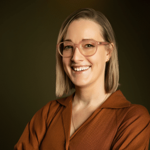 A woman with blonde hair and pink glasses smiles, wearing a brown collared shirt, standing against a plain dark background.