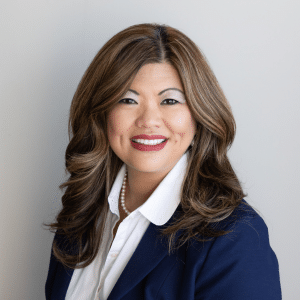 Woman with long, wavy brown hair wearing a navy blazer, white blouse, and pearl necklace, smiling in front of a plain light gray background.