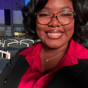 A woman wearing glasses, a red blouse, and a black blazer smiles outdoors with rows of white chairs and a modern building in the background, illuminated by sunlight.