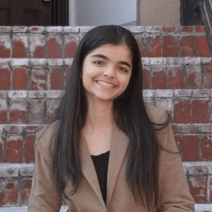 A young woman with long dark hair, wearing a brown blazer and black top, smiles while sitting on outdoor brick steps.