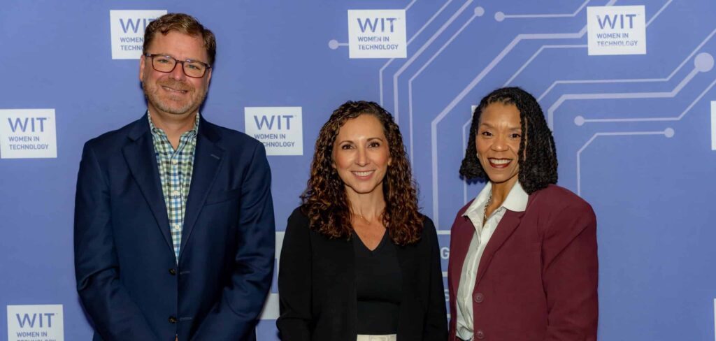 Three people stand and smile in front of a blue Women in Technology (WIT) backdrop at an event. Two women and one man are dressed in professional attire.