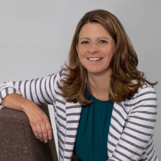 A woman with wavy brown hair, wearing a white and gray striped blazer over a teal top, sits on a brown chair, smiling at the camera against a plain light gray background.