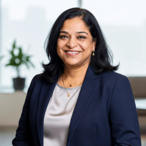 A woman with medium-length black hair, wearing a navy blazer and gray blouse, smiles in an office setting with a blurred plant and window in the background.