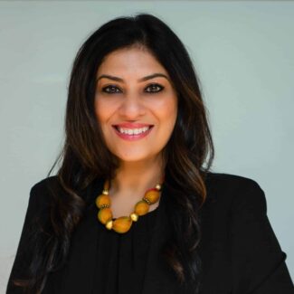 A woman with long dark hair wearing a black blazer, a chunky beaded necklace, and several bracelets, smiling and standing against a light gray background.