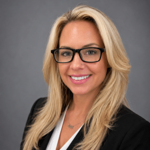 A woman with long blonde hair, black glasses, and a black blazer over a white blouse smiles in front of a plain gray background.