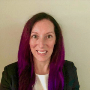 Woman with long straight dark purple hair wearing a black blazer and white top, smiling at the camera, with a plain light-colored background.
