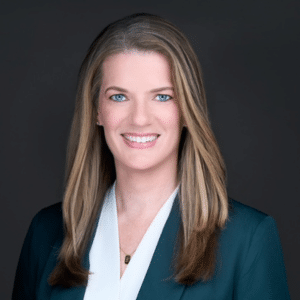Professional woman with long, straight, light brown hair wearing a dark blazer over a white blouse, smiling at the camera against a dark background.