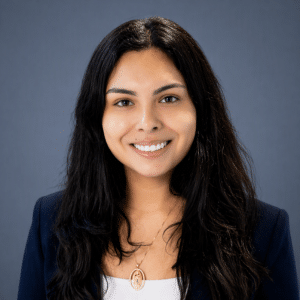 A woman with long dark hair, wearing a navy blazer, white top, and a necklace, smiles at the camera against a plain blue-gray background.