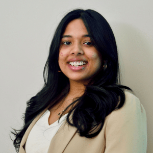A young woman with long black hair, wearing a beige blazer and white top, smiles at the camera against a plain light background.